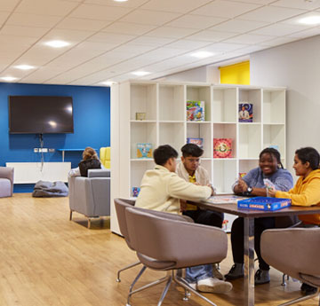 Four students sitting round their kitchen table playing a game whilst another student sits on a sofa watching TV